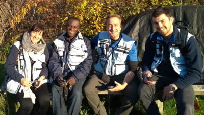 Four MapAction volunteers on a training exercise sitting outdoors in a row, smiling to camera.