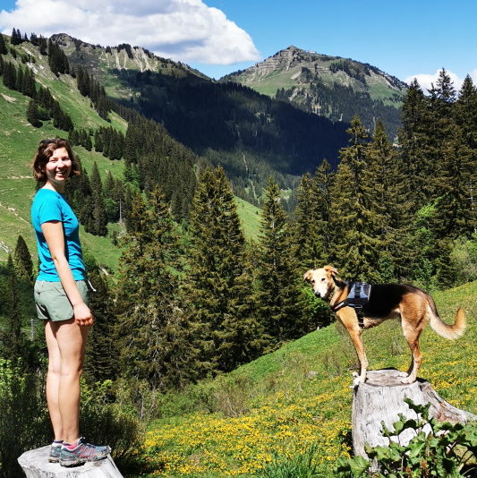 Emma Hall and her dog Woody each standing on a tree stump on a sunny Austrian mountainside, looking to camera.