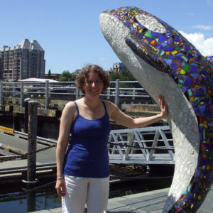 Gemma with her hand on a colourful statue of a leaping dolphin on a sunny day. She is smiling to camera.