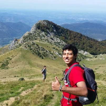 Daniel on a sunny mountain top wearing a backpack and smiling to camera. Another climber is visible in the background also looking to the camera.