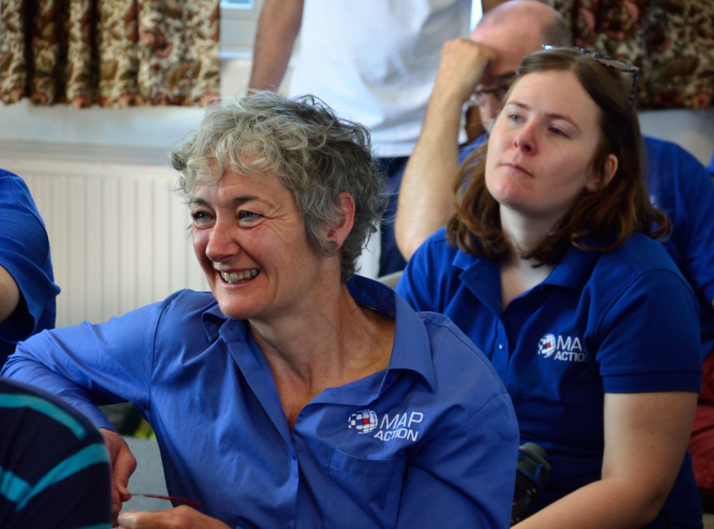 Picture of Liz Hughes smiling, seated among MapAction team members