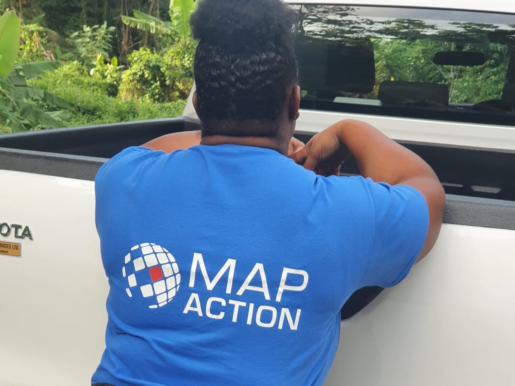 Lavern in the back of a truck driving through a rural tropical location, wearing a MapAction teeshirt with a large logo on her back.
