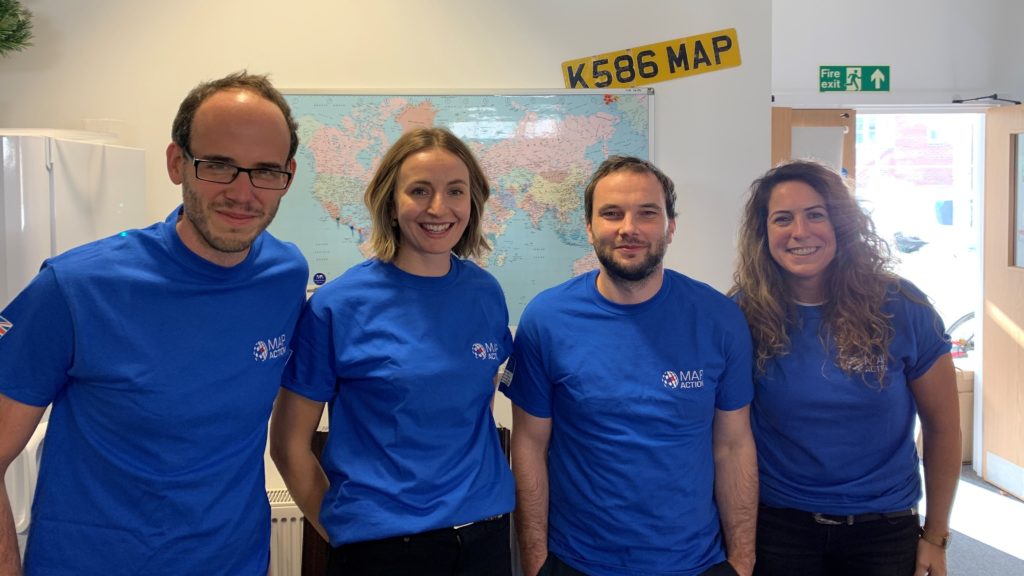2021 volunteer intake - four new volunteers with Mapction T-shirts smile at the camera in an office with a map behind them.